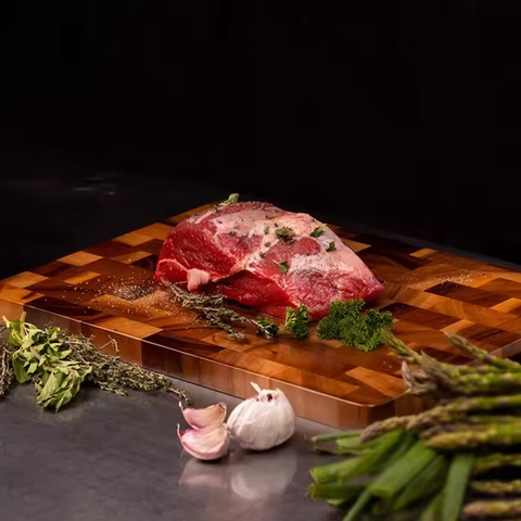 Raw meat on a wooden cutting board with herbs and garlic cloves on a dark background