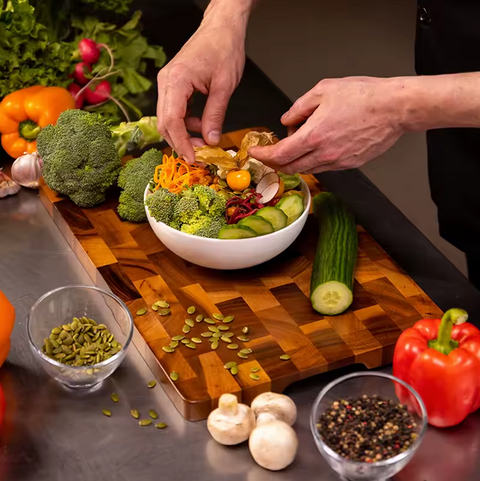 Person preparing a salad with various vegetables on a wooden acacia cutting board.